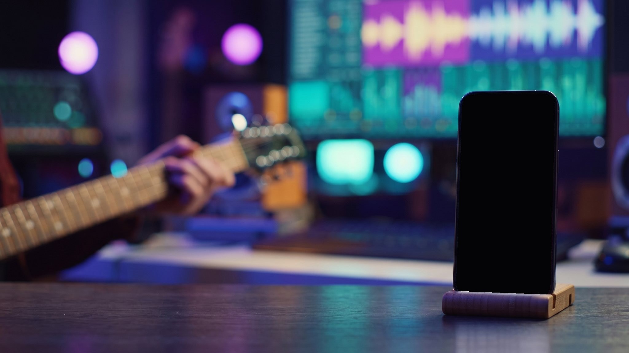 My Studio Pulse running on an iPhone in a wooden stand on a recording studio desk, with a guitarist and a mixing console out of focus behind it
